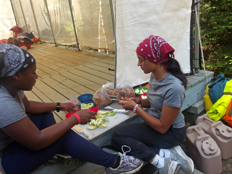 Two women are preparing food outdoors. One woman, wearing a red bandana, sits on a step and cuts vegetables on a cutting board. The other woman, wearing a black bandana, sits beside her and also cuts vegetables. They are in front of a tent-like structure with supplies nearby.